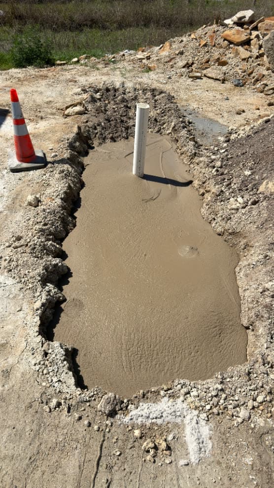 Freshly poured flowable fill filling a rectangular excavation, marked by a traffic cone